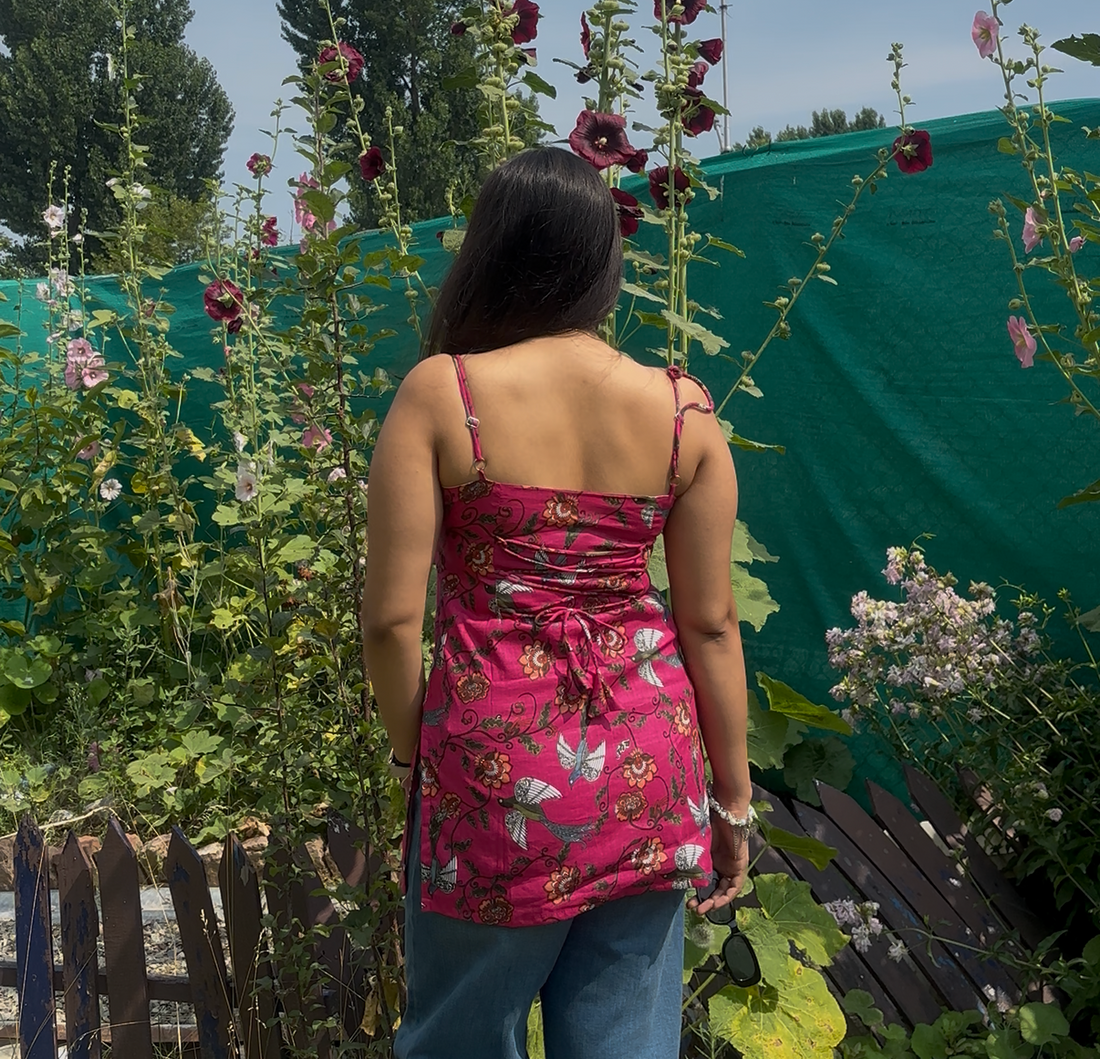 A woman viewed from behind wearing a pink floral spaghetti-strap top and blue jeans, standing in a garden with tall hollyhocks and a green tarp background.